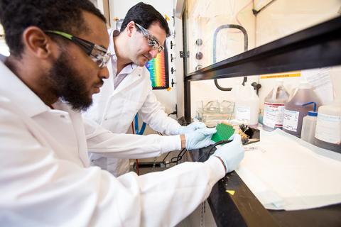 Two researchers in white coats and safety glasses study a circuit board