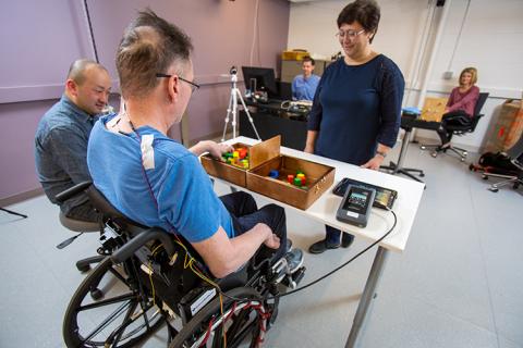 Spinal stimulation is applied by the stimulator (right) while video and EMG data are collected during the box-and-blocks motor skill task.