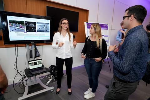 A young woman standing in front of a tv monitor, explaining her research to another young woman and young man