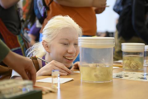 A smiling young woman holds a pencil and crouches in front of a jar with a brain