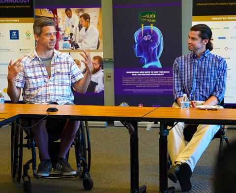 Man in wheelchair on left, sitting next to man on right, both behind a table, speaking to an audience
