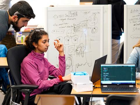 A male student looking over the shoulder of a female student who is working on a laptop with calculations on a board behind them.