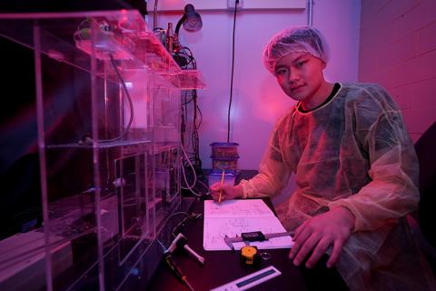 A young man in sterile lab clothing sits at a desk in a darkened lab.