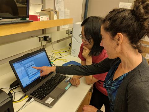 A woman points to a computer screen in a lab setting with a young woman standing next to her, looking at the screen