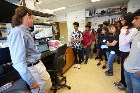 A group of students in a lab, listening to a male instructor, who is standing next to a monitor