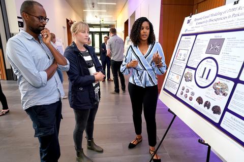 A young woman standing next to a research poster, explaining her work to a young man and another young woman standing nearby