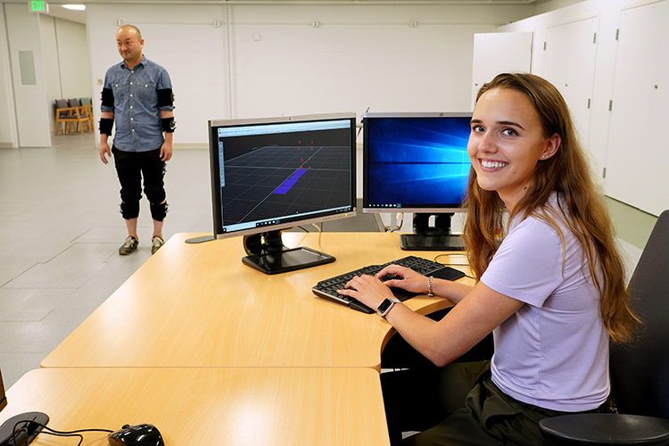 A young woman sits at a desk monitoring a man covered with sensors who is walking in the background