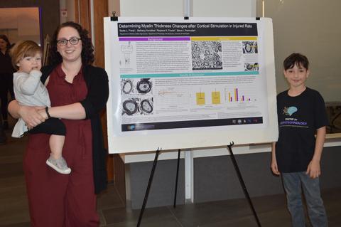 The RET program culminates in an end-of-summer research symposium where participants explain their research project in front of an audience and display a research poster they develop. RET participant, Sadie Frady, stands next to her research poster with her children.